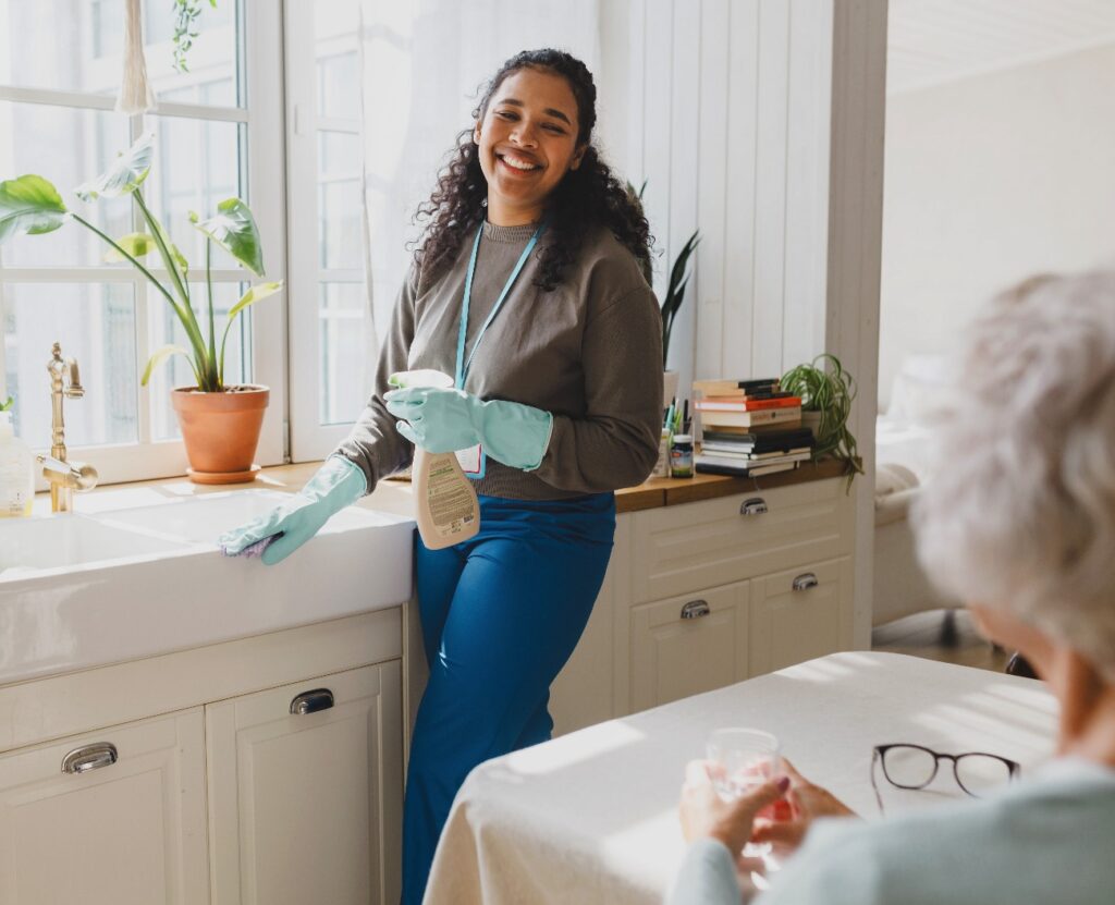 Der Fokus liegt auf einer fröhlichen afroamerikanischen Freiwilligen, die einer älteren Dame bei der Hausarbeit hilft; sie steht in der Küche in rosafarbenen Handschuhen, wäscht das Spülbecken mit Spülmittel und unterhält sich mit der älteren Dame, die am Tisch sitzt.