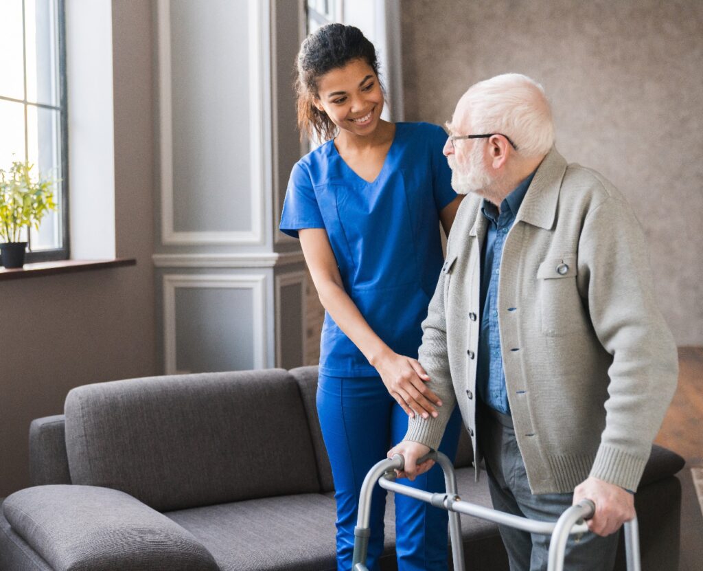Porträt einer jungen afrikanischen Pflegekraft, die einem älteren, behinderten Mann – ihrem Großvater – im Schlafzimmer dabei hilft, mit einem Rollator zu gehen. Ein älterer Patient eines Pflegeheims bewegt sich mit Hilfe eines Rollators und der Unterstützung einer Pflegekraft fort.